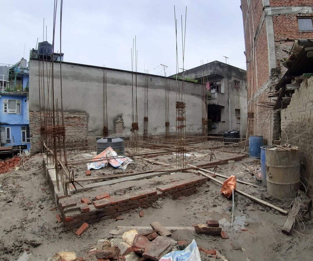 Foundation stage on a tight urban building site: vertical rebar column cages rising from brick footings and ground beams, with water tanks and supplies stacked beside neighbouring houses