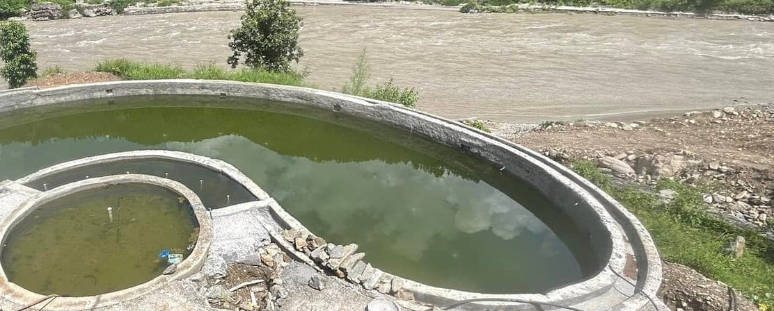 Curved concrete pool terrace under construction on a slope above a wide, fast-flowing river, with forested hills and village houses on the far bank